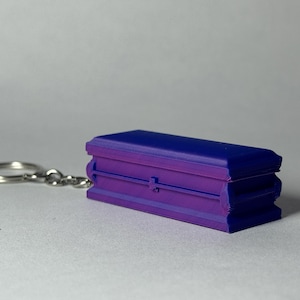 May include: A miniature coffin keychain in shades of purple and blue. The small coffin has a rectangular shape with a hinged lid and is attached to a silver keyring. The keychain is a novelty item.