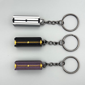 May include: Three rectangular keychains with metal rings. The top keychain is silver with a black center line. The middle keychain is black with a gold center line. The bottom keychain is dark purple with a gold center line. Each has a chain link.