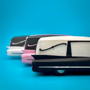 May include: Three miniature toy hearses arranged in a row, each featuring a distinct colour scheme. The front vehicle is black and white, the middle is pink and black, and the rear is white and black. Each model displays classic tail fins and side trim.
