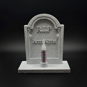 May include: A gray, textured, stone-like tombstone ornament featuring the embossed text R.I.P. and Aron Alpha. A small, clear plastic tube of Aron Alpha adhesive with a red warning label is positioned upright in a circular holder on the base.