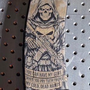 May include: A tan and black magazine featuring a hooded skull graphic holding a rifle. The text "YOU CAN HAVE MY GUNS WHEN YOU PRY THEM FROM MY COLD, DEAD HANDS" is below the graphic. The magazine is for a .56 x 45 calibre weapon.