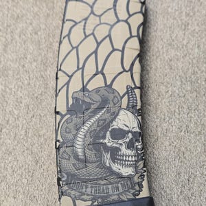 May include: A tan rifle magazine featuring a black graphic print of a coiled rattlesnake wrapped around a human skull. The design includes a scale-like pattern on the upper portion and the text "DONT TREAD ON ME" printed on a banner below the skull.