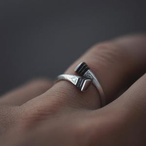 Puede incluir: Un anillo de plata con un diseño único. El anillo presenta dos formas geométricas angulares que se encuentran en el dedo. Cada forma tiene una serie de líneas oscuras. El anillo se muestra en un dedo, con un fondo borroso.