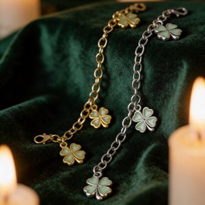 May include: Two charm bracelets with four-leaf clover pendants. One bracelet is gold-toned, the other is silver-toned. The clovers have a light green enamel fill. The bracelets are displayed on a dark green velvet surface, with lit candles in the background.