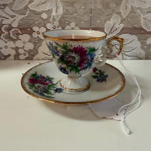 May include: A vintage teacup candle with floral designs in pink, blue, and green. The teacup sits on a matching saucer, both with gold trim. A white candle is inside the teacup.