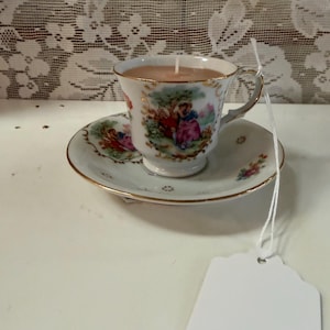 May include: A vintage teacup candle set on a matching saucer. The teacup and saucer are white with a gold trim and a pastoral scene. The candle wax is light brown, and a white wick is visible. A blank tag hangs nearby.