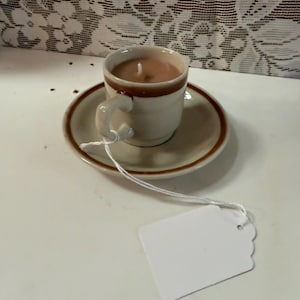 May include: A small, beige teacup candle with a brown rim sits on a matching saucer. A white tag with a string is attached to the cup. The candle has a light brown wax and a white wick.