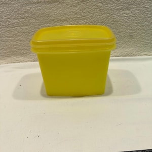 May include: A bright yellow, square-shaped plastic container with a matching lid. The container has a slightly rounded top and a smooth, matte finish. The lid fits snugly on top, creating a sealed appearance. The container is empty.