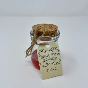 Puede incluir: Un pequeño frasco de vidrio con un tapón de corcho y una cuerda. El frasco contiene un líquido rojo y tiene una etiqueta que dice "Regular Potion of Healing" y "2D4+2". Sobre un fondo blanco.