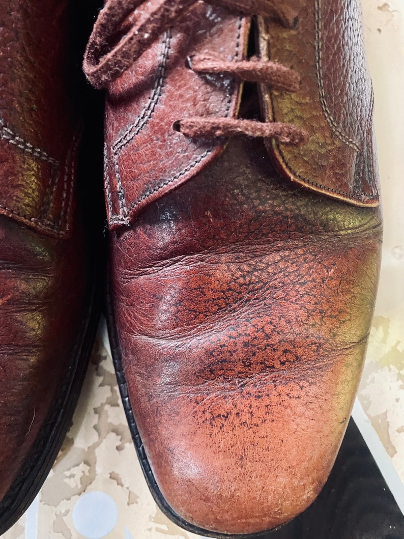 May include: Close-up of a pair of brown leather shoes with laces. The shoes have a worn and distressed look, with visible stitching and creases.