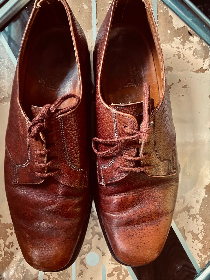 May include: A pair of brown leather lace-up shoes with a slightly rounded toe. The shoes have a worn look and are made of a textured leather.