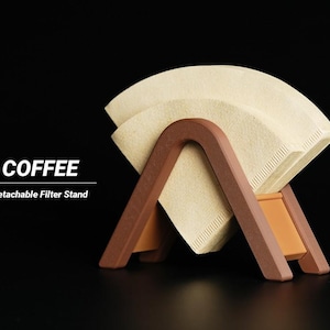 Può includere: Un supporto per filtro da caffè in legno marrone che contiene due filtri di carta beige. Il supporto ha un design unico a forma di A con una base rettangolare. La parola "COFFEE" e "Detachable Filter Stand" sono stampate.