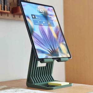 May include: A green tablet stand holding a tablet displaying a floral image and the time 11:43. The stand has a ribbed design and a small tray with a yellow sticky note pad. The tablet is angled upwards.