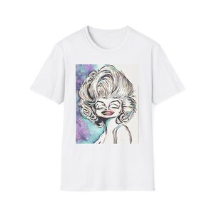 May include: White t-shirt featuring a colorful illustration of a smiling woman with voluminous blonde hair. The artwork has a cartoonish style with bold lines and a vibrant color palette, set against a purple and teal background.