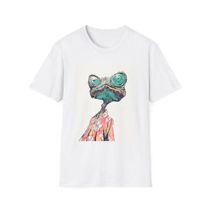 May include: White t-shirt featuring a whimsical illustration of a chameleon-like creature with large, expressive eyes. The creature is wearing a pink Hawaiian shirt with white flowers. The design is centered on the shirt.