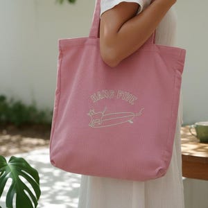 Könnte beinhalten: Eine rosa Cord-Einkaufstasche mit dem Schriftzug "HANG FIVE" und einem Dackel auf einem Surfbrett, weiß gestickt. Die Tasche hat zwei Schultergurte und eine rechteckige Form, ideal zum Tragen von Gegenständen.