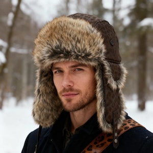 Pode incluir: Um chapéu de caçador de pele sintética castanho e bege com protetores de orelha. O chapéu tem um padrão de leopardo no exterior e é usado com um casaco azul escuro e uma alça com estampa de leopardo.