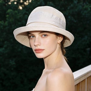 Puede incluir: Un sombrero para el sol color crema con ala ancha y corona estructurada. El sombrero tiene una textura sutil y un detalle de banda alrededor de la corona. El fondo es una vista borrosa de follaje verde y una terraza de madera.