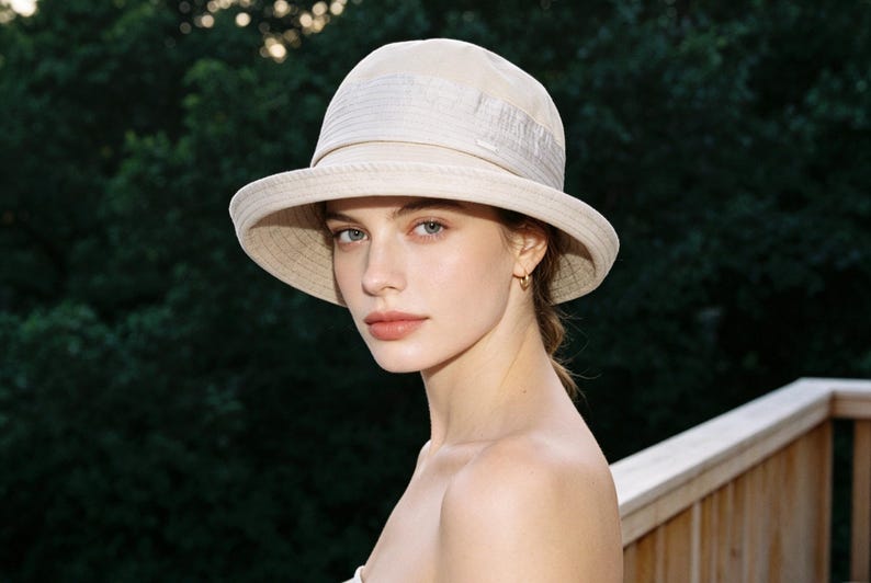Puede incluir: Un sombrero para el sol color crema con ala ancha y corona estructurada. El sombrero tiene una textura sutil y un detalle de banda alrededor de la corona. El fondo es una vista borrosa de follaje verde y una terraza de madera.
