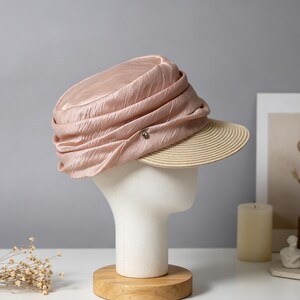 May include: A light pink hat with a pleated crown and a beige brim. The hat is displayed on a white mannequin head. A small silver button is on the side of the hat. The hat is a stylish accessory.
