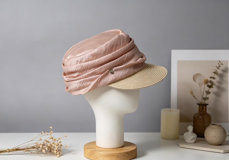 May include: A light pink hat with a pleated crown and a beige brim. The hat is displayed on a white mannequin head. A small silver button is on the side of the hat. The hat is a stylish accessory.