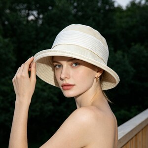 Puede incluir: Un sombrero para el sol color crema con ala ancha y una banda blanca. El sombrero est&aacute; hecho de un material tejido y tiene un dise&ntilde;o cl&aacute;sico y elegante. El fondo es una vista borrosa de &aacute;rboles verdes.