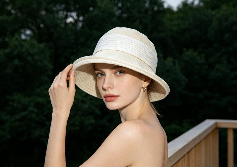Puede incluir: Un sombrero para el sol color crema con ala ancha y una banda blanca. El sombrero est&aacute; hecho de un material tejido y tiene un dise&ntilde;o cl&aacute;sico y elegante. El fondo es una vista borrosa de &aacute;rboles verdes.
