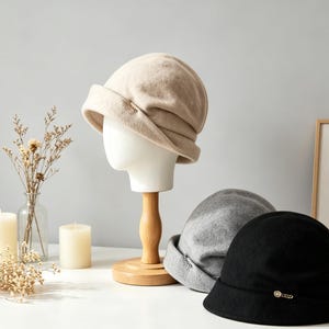 May include: Three wool cloche hats in beige, gray, and black are displayed on a white surface. The beige hat is on a mannequin head. Each hat has a small decorative accent. The hats are a classic style.