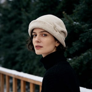 May include: A cream-coloured wool beret with a small gold chain detail. The beret is worn with a black turtleneck sweater. The background features a wooden railing and evergreen trees.