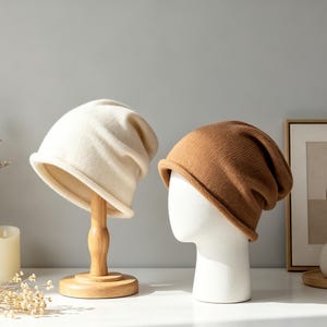 May include: Two knit beanies, one cream and one brown, displayed on mannequin heads. The cream beanie is on a wooden stand. The hats are on a white surface with a neutral background. Dried flowers and candles are in the background.