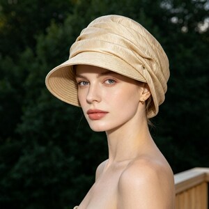 May include: A beige sun hat with a wide brim and a gathered crown. The hat has a structured design and a small gold-colored button detail. The hat is made of a woven material and is suitable for sun protection.