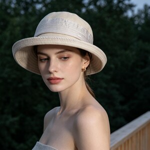 Puede incluir: Un sombrero para el sol color crema con ala ancha y detalle de cinta. El sombrero lo lleva una persona de piel clara y cabello casta&ntilde;o claro. El fondo es una vista borrosa de &aacute;rboles verdes.