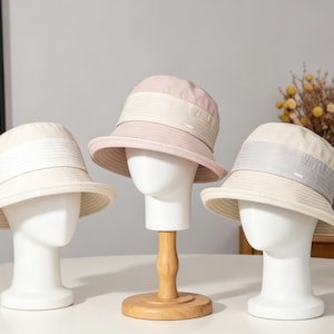 Puede incluir: Tres sombreros de pescador en crema, rosa y gris, exhibidos en cabezas de maniqu&iacute; blancas. Los sombreros tienen un ala ancha y una banda en contraste alrededor de la corona. Los sombreros est&aacute;n sobre una mesa blanca.