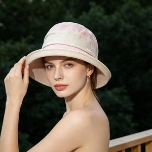 Puede incluir: Un sombrero para el sol rosa claro y beige con ala ancha. El sombrero tiene una copa redondeada y una banda en contraste en la base. La mujer sostiene el ala del sombrero.