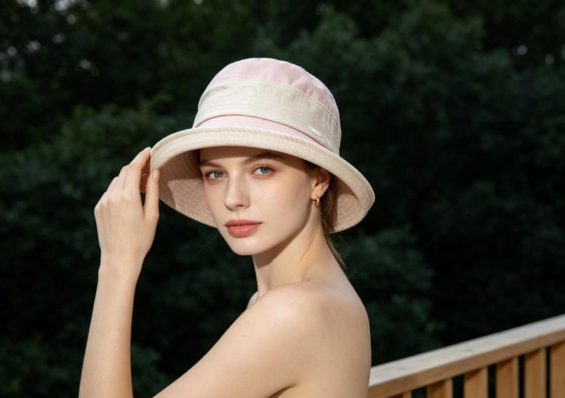 Puede incluir: Un sombrero para el sol rosa claro y beige con ala ancha. El sombrero tiene una copa redondeada y una banda en contraste en la base. La mujer sostiene el ala del sombrero.