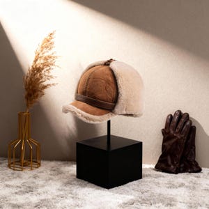 Könnte beinhalten: Ein brauner und beiger Winterhut mit Lederriemen und Ohrenklappen. Der Hut wird auf einem schwarzen Ständer neben einem Paar brauner Lederhandschuhe ausgestellt. Links befindet sich eine goldene Vase mit getrocknetem Gras.
