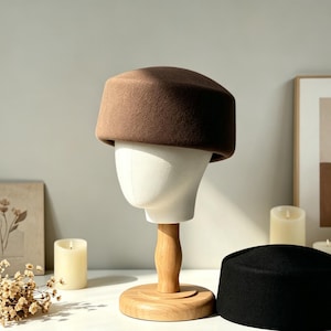May include: A brown felt hat sits atop a white mannequin head on a wooden stand. A black felt hat rests on a white surface. The hats have a rounded, flat-top design. The scene includes candles, a framed print, and a vase with greenery.