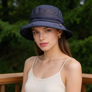 May include: A navy blue bucket hat with a mesh crown and a wide brim. The hat features a dark ribbon and a small gold-colored button. The hat is worn by a person with long brown hair and gold hoop earrings.