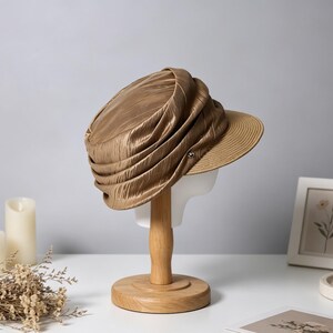 May include: A gold-colored hat with a pleated design and a brim, displayed on a wooden stand. The hat has a textured appearance and a small button detail. The brim is a lighter shade of brown. The hat is on a white mannequin head.