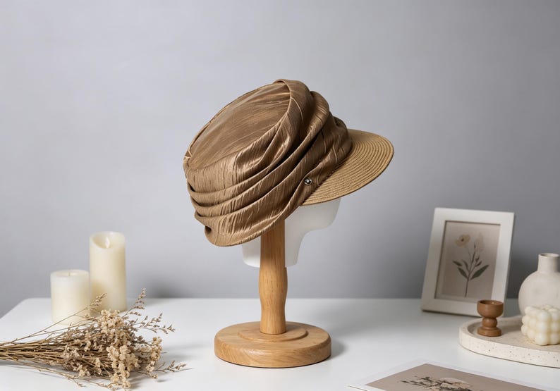 May include: A gold-colored hat with a pleated design and a brim, displayed on a wooden stand. The hat has a textured appearance and a small button detail. The brim is a lighter shade of brown. The hat is on a white mannequin head.