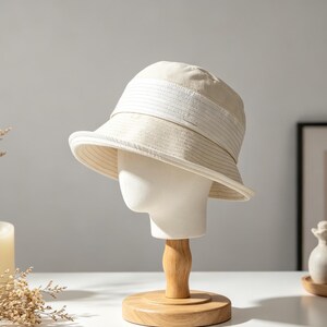 Puede incluir: Un sombrero de pescador beige con un detalle de banda blanca se exhibe en una cabeza de maniqu&iacute;. El sombrero est&aacute; hecho de una tela texturizada y tiene un ala ancha. El maniqu&iacute; est&aacute; sobre un soporte de madera.