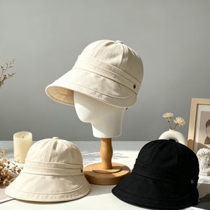 May include: Three hats in a studio setting. A cream-colored hat is displayed on a mannequin head. Two additional hats are on a white surface: one cream-colored and one black. All hats have a similar design with a brim and a band.