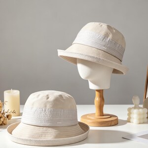 Puede incluir: Dos sombreros de cubo beige con un detalle de banda de color claro. Un sombrero se exhibe en una cabeza de maniqu&iacute; con un soporte de madera. El otro sombrero se coloca sobre una superficie blanca. Los sombreros son parte de una colecci&oacute;n de accesorios de moda.