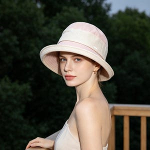 Puede incluir: Un sombrero para el sol de color rosa claro con ala ancha y una banda color crema. El sombrero tiene una copa redondeada. El fondo es una vista borrosa de &aacute;rboles verdes y una terraza de madera. El sombrero se usa al aire libre.