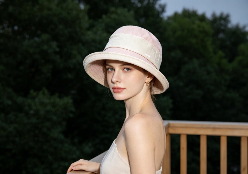 Puede incluir: Un sombrero para el sol de color rosa claro con ala ancha y una banda color crema. El sombrero tiene una copa redondeada. El fondo es una vista borrosa de &aacute;rboles verdes y una terraza de madera. El sombrero se usa al aire libre.