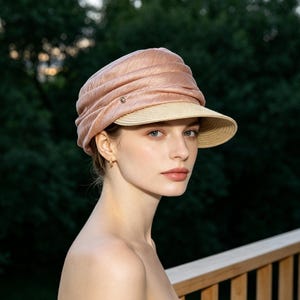 May include: A blush-colored hat with a structured, pleated crown and a beige brim. The hat features a small, decorative element on the side. The hat is designed for sun protection and fashion.