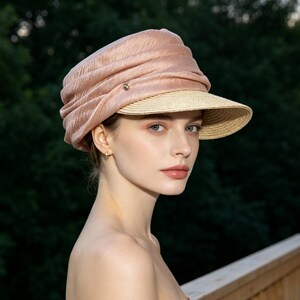 May include: A blush-colored hat with a woven brim and a gathered fabric crown. The hat features a small, gold-toned button detail. The hat is worn by a person with fair skin and blue eyes, against a backdrop of green foliage.