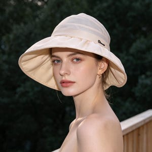 May include: A cream-colored sun hat with a wide brim. The hat has a slightly rounded crown and a ribbon detail. The brim is gently curved, providing shade. The hat is worn outdoors, with a blurred green background.