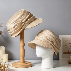 May include: Two beige hats with a textured design. One hat is displayed on a wooden hat stand, the other on a white mannequin head. The hats have a small brim and a decorative element on the side.