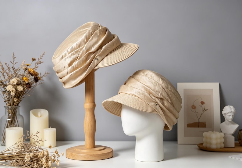 May include: Two beige hats with a textured design. One hat is displayed on a wooden hat stand, the other on a white mannequin head. The hats have a small brim and a decorative element on the side.